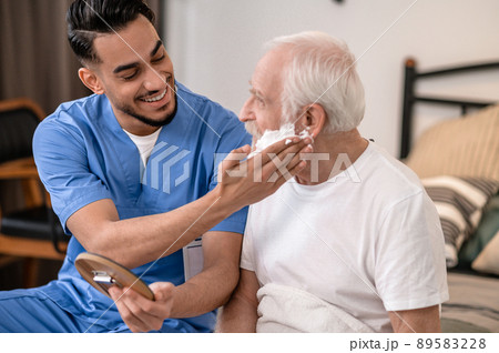 Joyful young volunteer preparing an old man for a shave Joyful young volunteer preparing an old man for a shave 89583228
