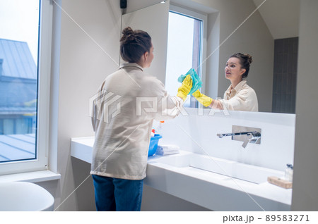 Cute young woman cleaning mirror in the bathroom Cute young woman cleaning mirror in the bathroom 89583271