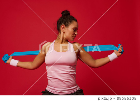 African American muscular woman doing stretching exercises on arms using elastic fitness band against red background with copy ad space 89586308