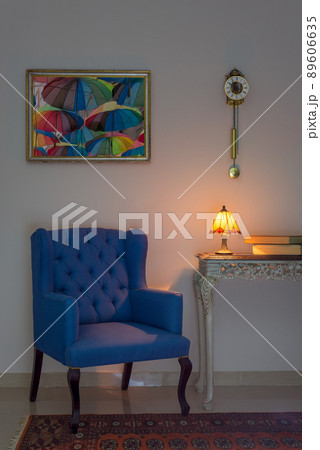 Interior composition of blue armchair, vintage wooden beige table, illuminated table lamp, books, pendulum clock and orange ornate carpet 89606635