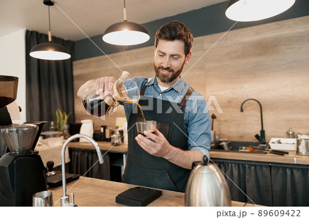Smiling barista with stylish beard wearing apron poured filter coffee into a glass Smiling barista with stylish beard wearing apron poured filter coffee into a glass 89609422