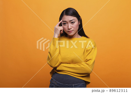 Serious pensive woman focused on introspection thoughts in a contemplation state on orange background. Concentrated young adult person in foreground standing and thinking about useful ideas. Serious pensive woman focused on introspection thoughts in a contemplation state on orange background. Concentrated young adult person in foreground standing and thinking about useful ideas. 89612311