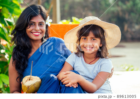 happy daughter in straw hat with her beautiful mother in blue saree drinking fresh coconut water in tropical paradise beach in Goa. using eco metal recyclable tube 89613005