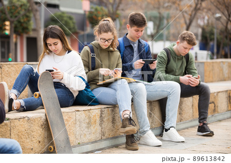 Four teenagers enthusiastically look at the screens of their smartphones on street Four teenagers enthusiastically look at the screens of their smartphones on street 89613842