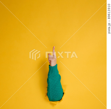 female hand sticks out of a torn hole in a yellow paper background, part of the body points with the index finger to top. Copy space female hand sticks out of a torn hole in a yellow paper background, part of the body points with the index finger to top. Copy space 89613910