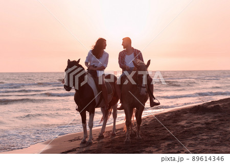 a loving young couple in summer clothes riding a horse on a sandy beach at sunset. Sea and sunset in the background. Selective focus  89614346