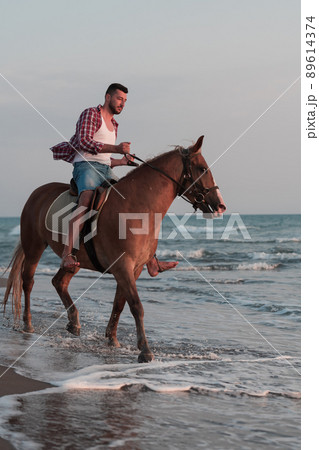 A modern man in summer clothes enjoys riding a horse on a beautiful sandy beach at sunset. Selective focus  89614374