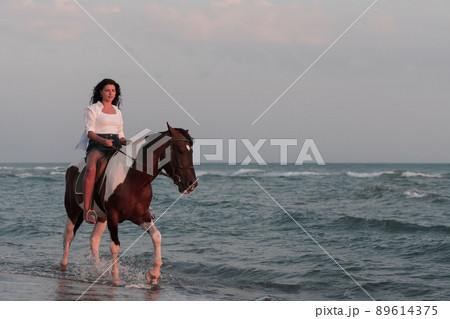 Woman in summer clothes enjoys riding a horse on a beautiful sandy beach at sunset. Selective focus Woman in summer clothes enjoys riding a horse on a beautiful sandy beach at sunset. Selective focus 89614375