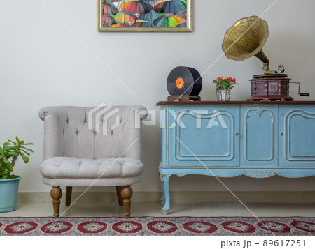 Retro off white armchair, vintage wooden light blue sideboard, old phonograph (gramophone) and vinyl records on background of beige wall, tiled porcelain floor, and red carpet Retro off white armchair, vintage wooden light blue sideboard, old phonograph (gramophone) and vinyl records on background of beige wall, tiled porcelain floor, and red carpet 89617251