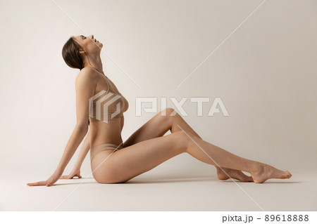 Side view portrait of young slim woman sitting on floor, posing in beige underwear isolated over grey studio background. Depilation, epilation concept 89618888