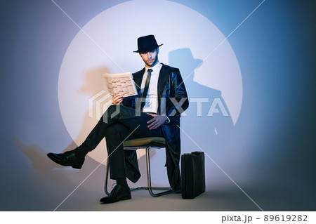 Handsome private detective sitting on chair reading newspaper in the spotlight on studio background Handsome private detective sitting on chair reading newspaper in the spotlight on studio background 89619282