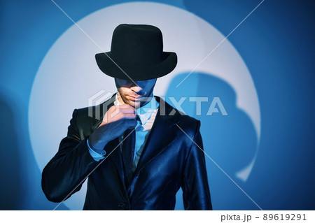 Portrait of handsome man in black coat hiding face behind his hat in the spotlight on studio background 89619291