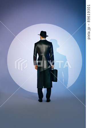 Back view of man silhouette in black coat and hat holding briefcase in the spotlight on studio background 89619301