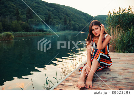 A young pretty woman poses sitting on a wooden pier by the river. Summer holiday 89623293