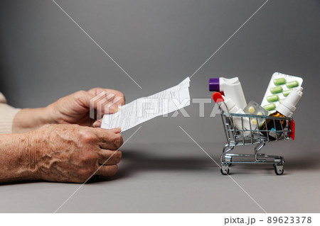 Close up of hands of senior woman holding an instructions of medicines near shopping cart. Gray background. Copy space. Concept of shopping pills and treatment Close up of hands of senior woman holding an instructions of medicines near shopping cart. Gray background. Copy space. Concept of shopping pills and treatment 89623378