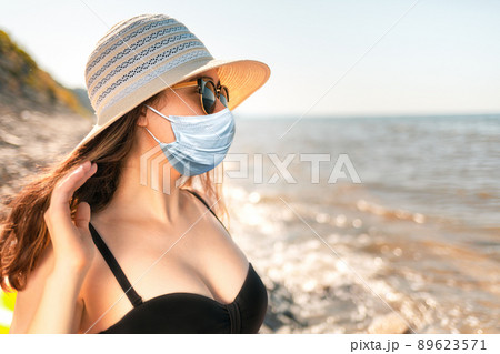 Portrait of a beautiful young woman, with full Breasts, in a bathing suit, wearing a hat and a medical mask. Beach and sea, windy weather. The concept of protection from the virus while on vacation 89623571