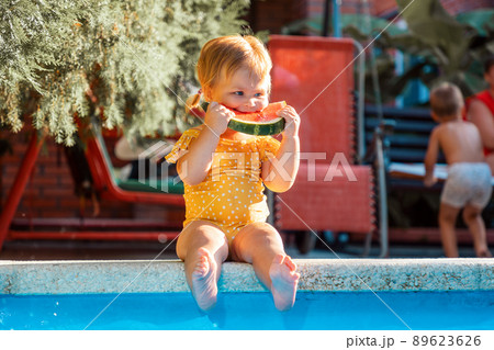 Summertime. Cute baby girl eating watermelon sitting by the pool. Concept of children's day Summertime. Cute baby girl eating watermelon sitting by the pool. Concept of children's day 89623626