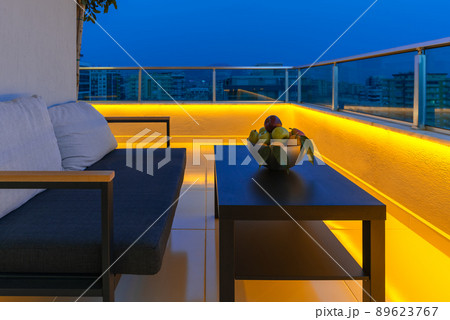 View of a cozy outdoor terrace with an armchair and a coffee table, LED lighting in the evening. View of a cozy outdoor terrace with an armchair and a coffee table, LED lighting in the evening. 89623767