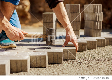 Young man laying gray concrete paving slabs in house courtyard on gravel foundation base. Master lays paving stones. Garden brick pathway paving by professional paver worker. Repairing sidewalk. Young man laying gray concrete paving slabs in house courtyard on gravel foundation base. Master lays paving stones. Garden brick pathway paving by professional paver worker. Repairing sidewalk. 89624030