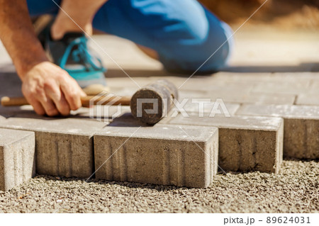 Young man laying gray concrete paving slabs in house courtyard on gravel foundation base. Master lays paving stones. Garden brick pathway paving by professional paver worker. Repairing sidewalk. 89624031