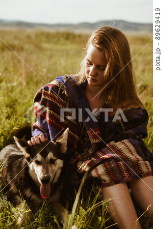 on the steppe grass against the background of the hills sits a girl in an ethno cape and strokes a dog on the steppe grass against the background of the hills sits a girl in an ethno cape and strokes a dog 89629419