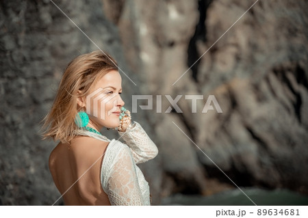 Middle aged woman looks good with blond hair, boho style in white long dress on the beach decorations on her neck and arms. 89634681
