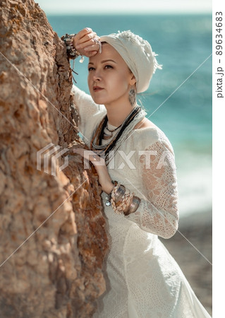 Middle aged woman looks good with blond hair, boho style in white long dress on the beach decorations on her neck and arms. 89634683