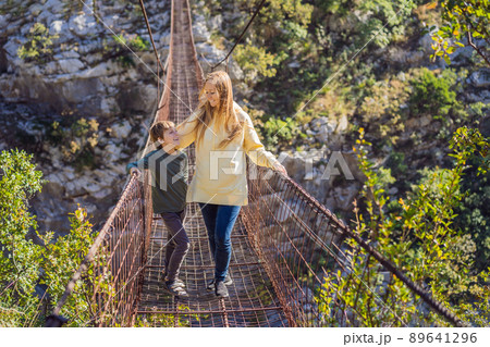 Mom and son tourists on Old rusty bridge. Attraction Long extreme suspension iron bridge across the river Moraca. Sights of Montenegro. Landmark Montenegro Mom and son tourists on Old rusty bridge. Attraction Long extreme suspension iron bridge across the river Moraca. Sights of Montenegro. Landmark Montenegro 89641296