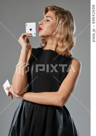 Blonde girl in black stylish dress showing two playing cards, posing against gray background. Gambling entertainment, poker, casino. Close-up. Blonde girl in black stylish dress showing two playing cards, posing against gray background. Gambling entertainment, poker, casino. Close-up. 89642698