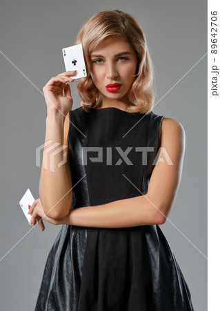 Blonde girl in black stylish dress showing two playing cards, posing against gray background. Gambling entertainment, poker, casino. Close-up. 89642706