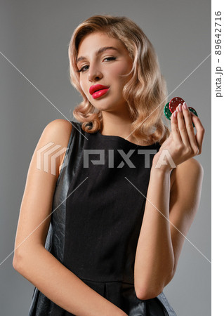 Blonde girl in black leather dress holding some colorful chips, posing against gray background. Gambling entertainment, poker, casino. Close-up. Blonde girl in black leather dress holding some colorful chips, posing against gray background. Gambling entertainment, poker, casino. Close-up. 89642716