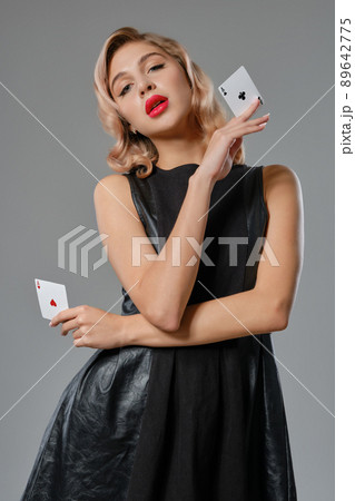 Blonde girl in black leather dress showing two playing cards, posing against gray background. Gambling entertainment, poker, casino. Close-up. 89642775