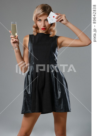 Blonde girl in black dress holding two playing cards and glass of champagne, posing against gray background. Gambling, poker, casino. Close-up. 89642838
