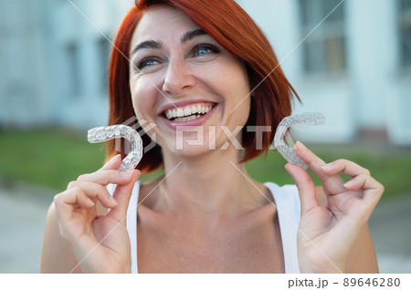 Red-haired Caucasian woman holding transparent mouthguards for bite correction outdoors. A girl with a beautiful snow-white smile uses silicone braces 89646280