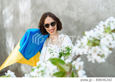 A girl in white dress and sunglasses in city on background of a concrete wall waves the national Ukrainian flag, celebrating victory. Apple flowers. Copy space. Conceptual idea - with Ukraine in heart A girl in white dress and sunglasses in city on background of a concrete wall waves the national Ukrainian flag, celebrating victory. Apple flowers. Copy space. Conceptual idea - with Ukraine in heart 89646282