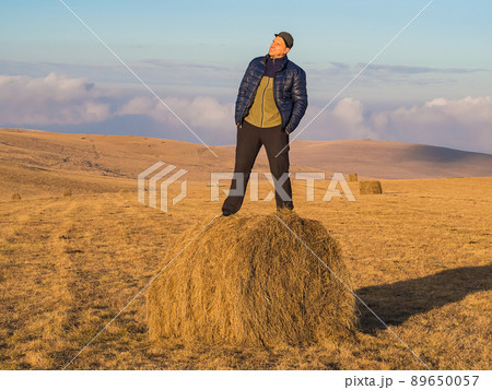 A man admires the picturesque surroundings while standing on a haystack in a wide golden field located in the Caucasian highlands. 89650057