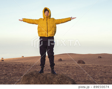 A cheerful rural girl stands with her hands apart on a haystack in a highland field under the sky A cheerful rural girl stands with her hands apart on a haystack in a highland field under the sky 89650074