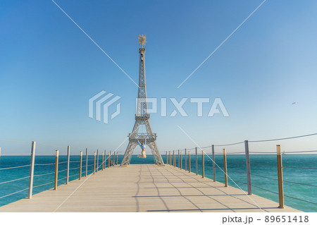 Large model of the Eiffel Tower on the beach. A woman walks along the pier towards the tower, wearing a blue jacket and white jeans. Large model of the Eiffel Tower on the beach. A woman walks along the pier towards the tower, wearing a blue jacket and white jeans. 89651418