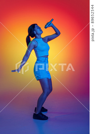 Emotive young girl singing in microphone, performing isolated over multicolored background in neon light 89652544