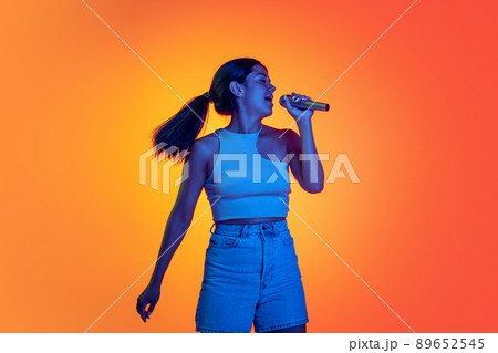 Half-length portrait of young girl singing in microphone isolated over orange background in neon light. Hobby 89652545