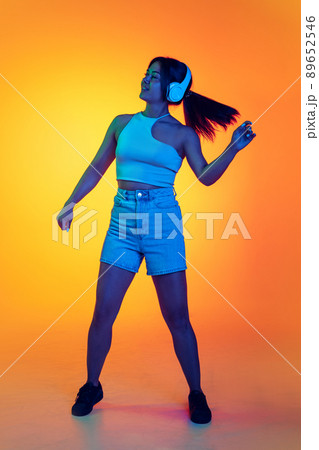 Full-length portrait of young girl listening to music in headphones, dancing isolated over orange background in neon light 89652546