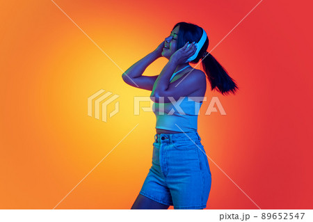 Young cheerful woman listening to music in headphones isolated over gradient yellow orange background in neon light Young cheerful woman listening to music in headphones isolated over gradient yellow orange background in neon light 89652547