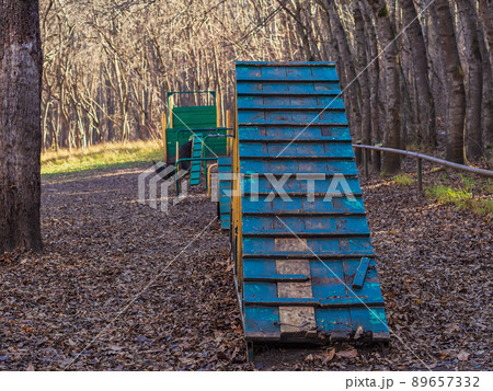 In the autumn forest in a clearing there is a training ground for dogs with wooden and metal barriers with peeling paint and broken parts. 89657332