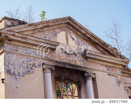 A fragment of an abandoned dilapidated and peeling Greek-style building overgrown with trees on the roof on a sunny day against the blue sky. 89657466