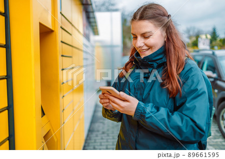 Caucasian woman showing smartphone with smart delivery app near a yellow automatic post terminal. Caucasian woman showing smartphone with smart delivery app near a yellow automatic post terminal. 89661595