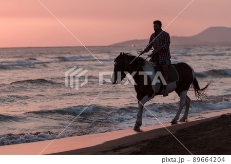 A modern man in summer clothes enjoys riding a horse on a beautiful sandy beach at sunset. Selective focus A modern man in summer clothes enjoys riding a horse on a beautiful sandy beach at sunset. Selective focus 89664204
