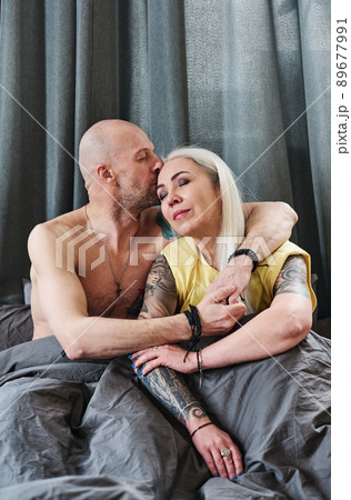 Portrait of modern mature Caucasian couple sitting on bed in morning, man kissing woman on forehead Portrait of modern mature Caucasian couple sitting on bed in morning, man kissing woman on forehead 89677991