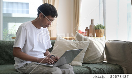 Man using laptop, sitting on couch at home, chatting or shopping in internet, reading message, working online with device. 89678487