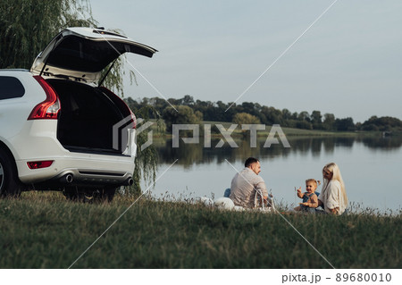 Happy Young Family Enjoying Picnic Time Outside the City, Mother and Father with Their Daughter Sitting Outdoors Near Their SUV Car, Road Trip Concept 89680010