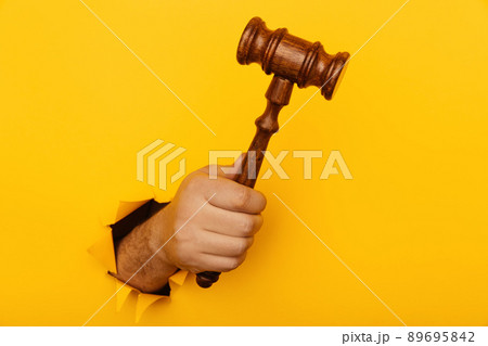 Hand holding a judge's gavel through torn yellow paper wall close-up. Law concept Hand holding a judge's gavel through torn yellow paper wall close-up. Law concept 89695842
